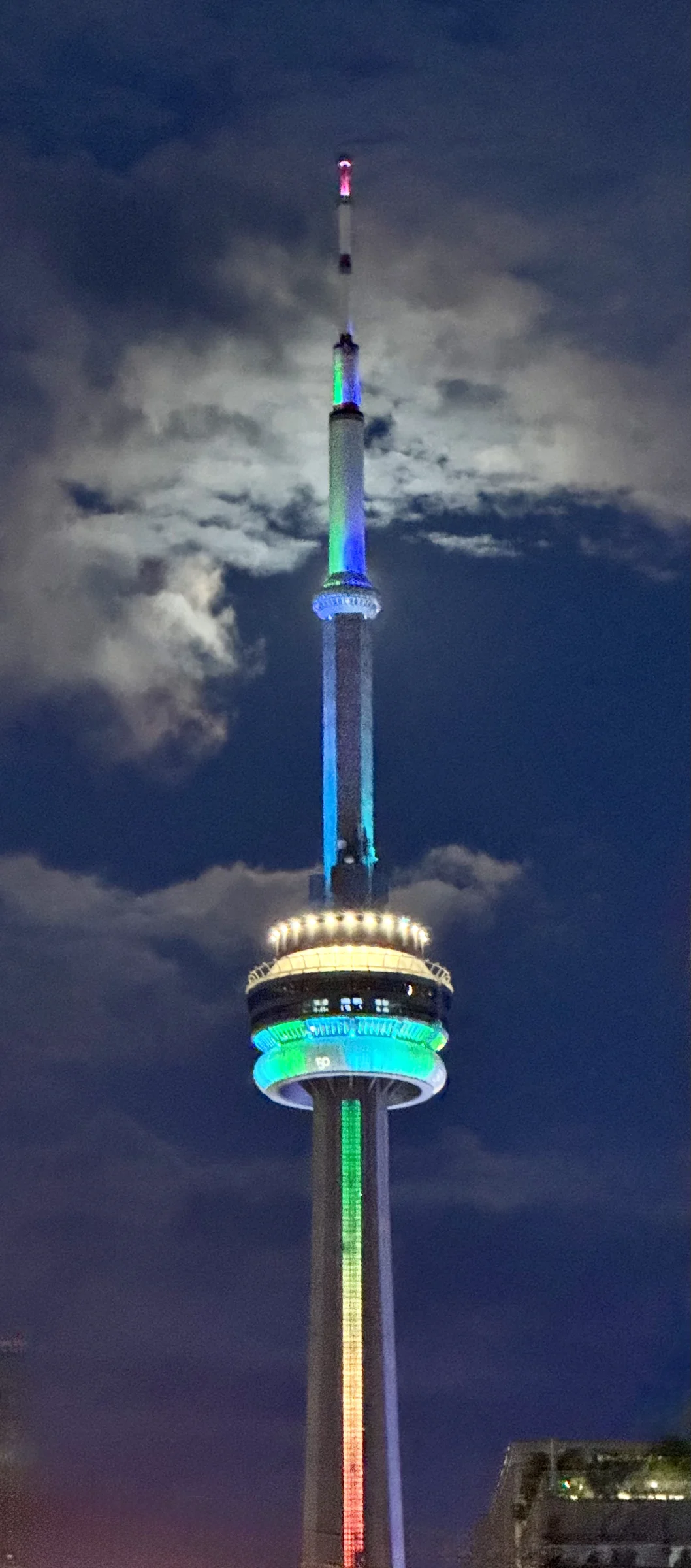 The CN Tower lit in rainbow colours at night, the moon half hidden behind its shaft