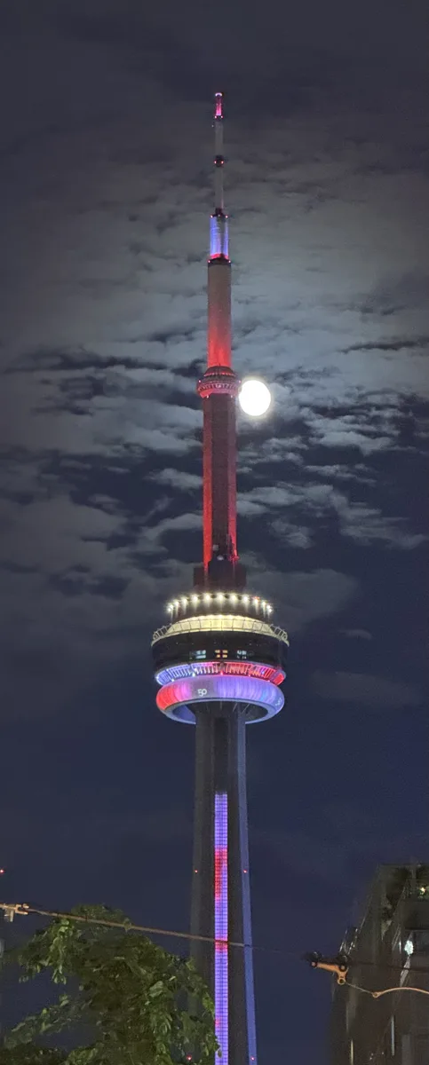 The CN Tower lit red and white at night with the full moon beside its shaft
